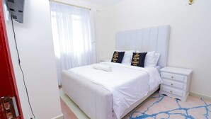 A hotel room with a bed, white curtains, a bedside table, and a rug. - White House (Nairobi)