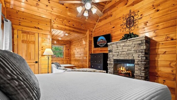 Ein gemütliches Blockhauszimmer mit Kamin aus Stein, einem Bett mit Kissen, einem Schreibtisch und einem Deckenventilator.