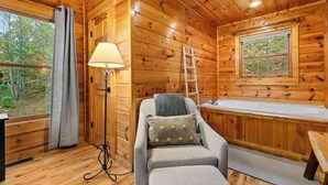 Ein gemütliches Blockhaus-Interieur mit Holzausstattung, einer Couch mit Kissen, einer Stehlampe, einer Leiter und einem Fenster mit Blick auf den Wald.