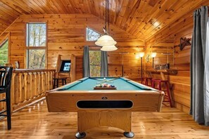 A wooden interior with a pool table, a bar area, and a window overlooking trees.