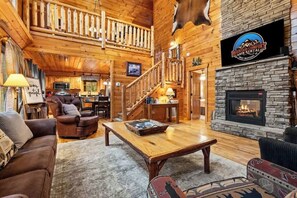 A cabin interior with a fireplace, wooden walls, a staircase, and a sign for Mountain Comfort Cabin Rentals. - Luxury Cabin • Hot Tub • Game Loft • 2 Kings (Sevierville)