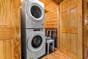 A stacked washer and dryer unit in a wooden cabin with a chair and a small table.