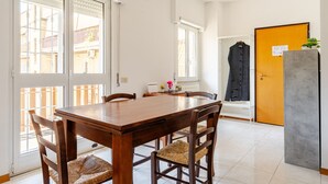 Een eethoek met een houten tafel en stoelen, een raam met zonwering, een radiator en een deur met een bordje.