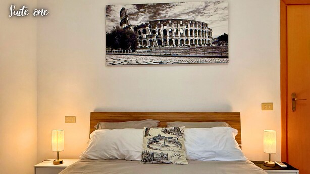 Een hotelkamer met een bed, twee nachtkastjes, een lamp, een aan de muur bevestigd schilderij van een historisch gebouw en een houten deur.
