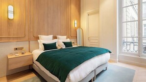 A modern bedroom with a large bed, wooden paneling on the walls, a nightstand, and a window with a view of buildings.