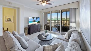 A modern living room with a large flat-screen TV, comfortable seating, a ceiling fan, and a balcony with a view of buildings and greenery.
