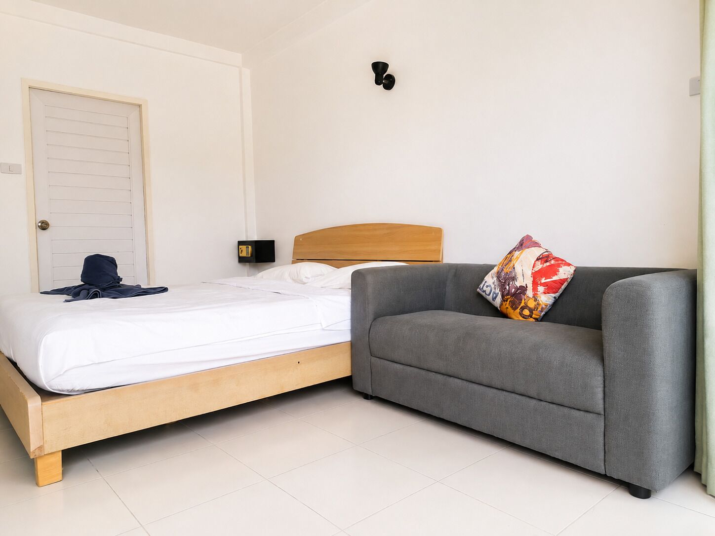 A bedroom with a bed, a grey sofa, a colorful pillow, a wall-mounted light, and a door.