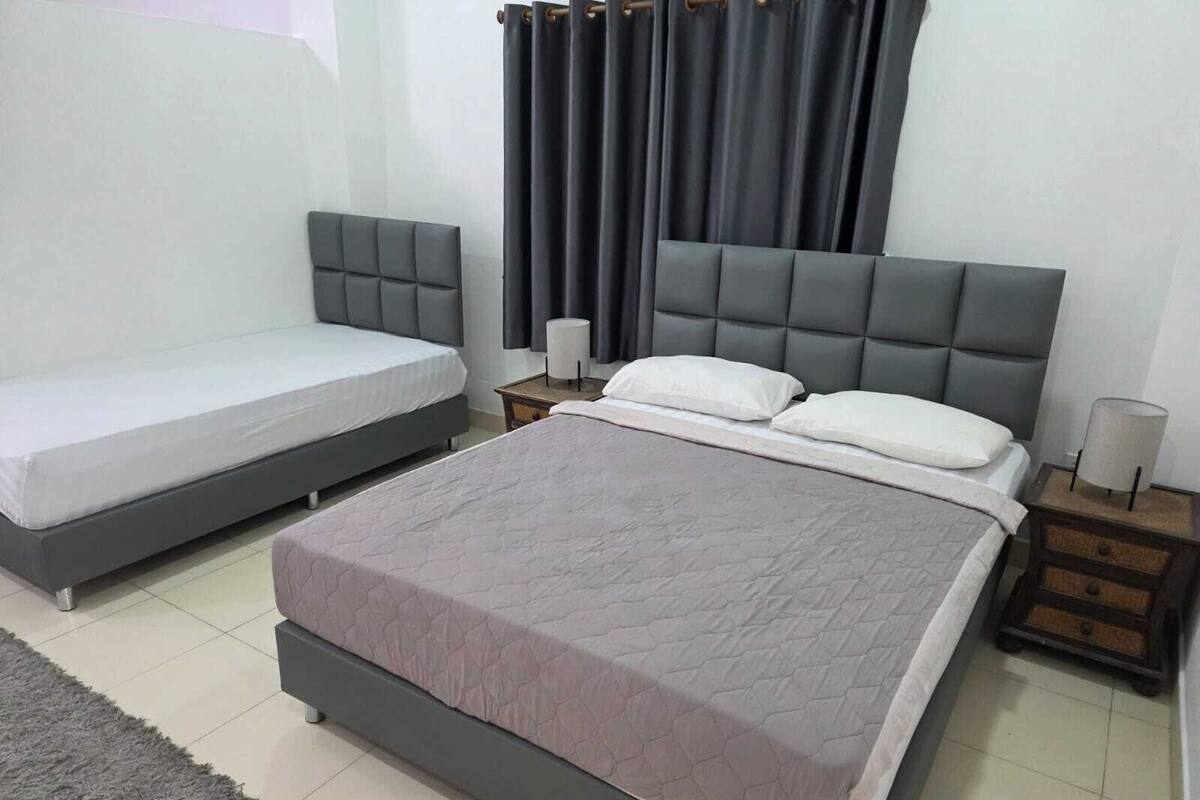 A bedroom with two beds, a grey headboard, a grey rug, and a nightstand with a lamp.