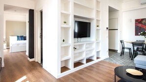 A modern living room with a TV on a white shelving unit, a dining area with a table and chairs, and a bedroom with a bed and nightstand.