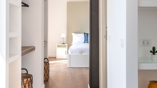 A hallway leading to a bedroom with a bed, bedside table, lamp, and a potted plant.