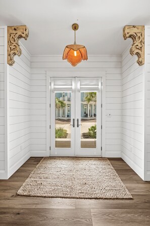 Una habitación con paredes blancas empapeladas, suelo de madera, alfombra tejida y una puerta que da a una casa azul.