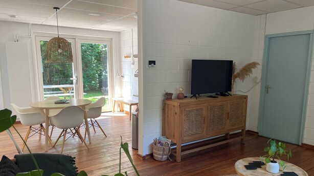 A modern living room with a wooden floor, a television on a wooden cabinet, a dining area with a table and chairs, and a large window with a view of greenery. - Seaside house in the heart of nature (Oosterend)