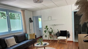 A modern living room with a black leather sofa, a round coffee table, and a wicker chair. There is a potted plant on a small table, and a large window provides natural light. - Seaside house in the heart of nature (Oosterend)