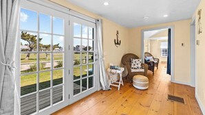 A room with a large sliding glass door leading to a garden, a wicker chair with cushions, a small table with a lamp, and a wooden floor. - Charming Seaside Cottage  Sleeps 9 with WiFi, AC, 2 Min Walk to a Private Beach (Sandwich)