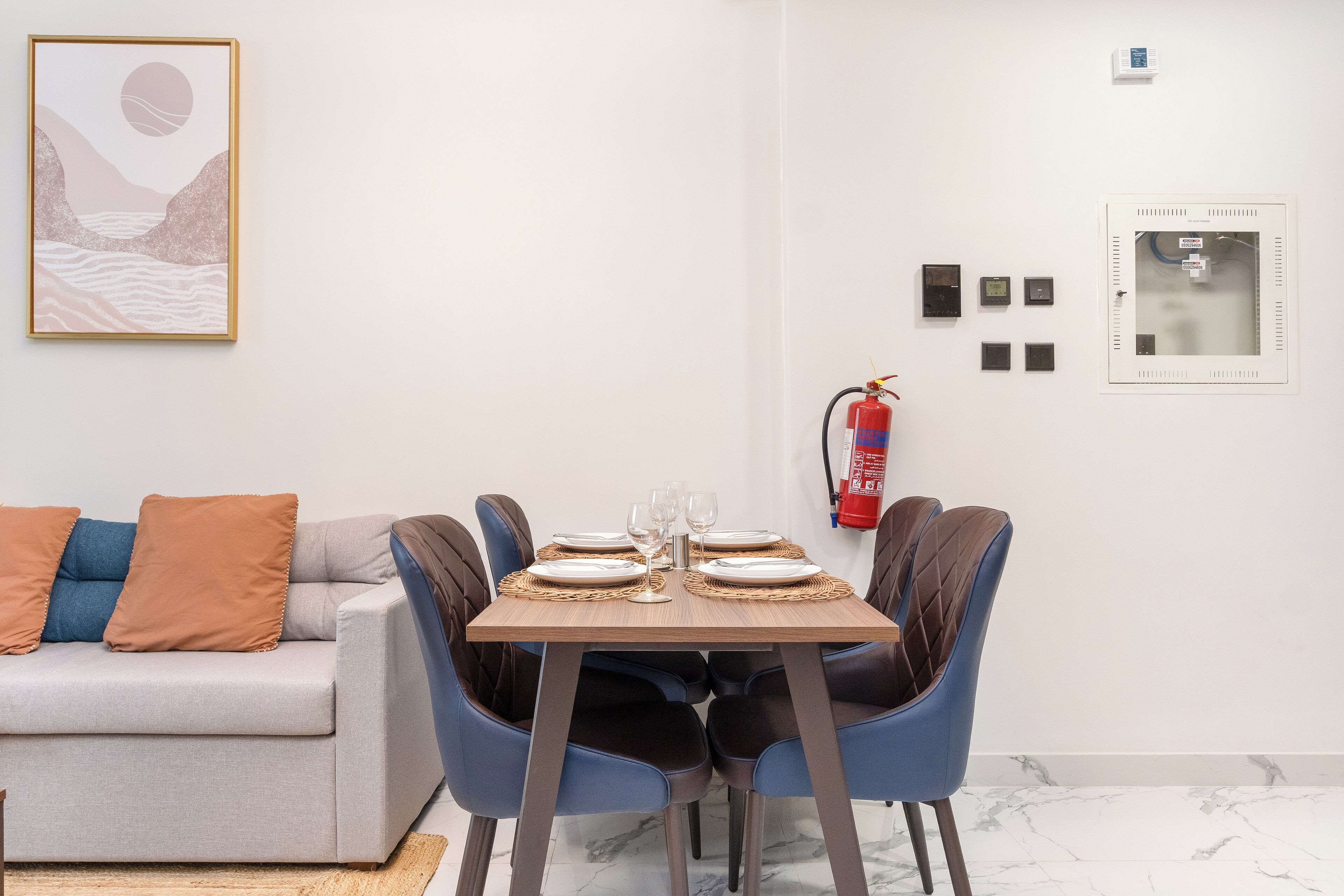 A dining area with a table set for four, blue chairs, a grey sofa with brown cushions, and a framed artwork on the wall.