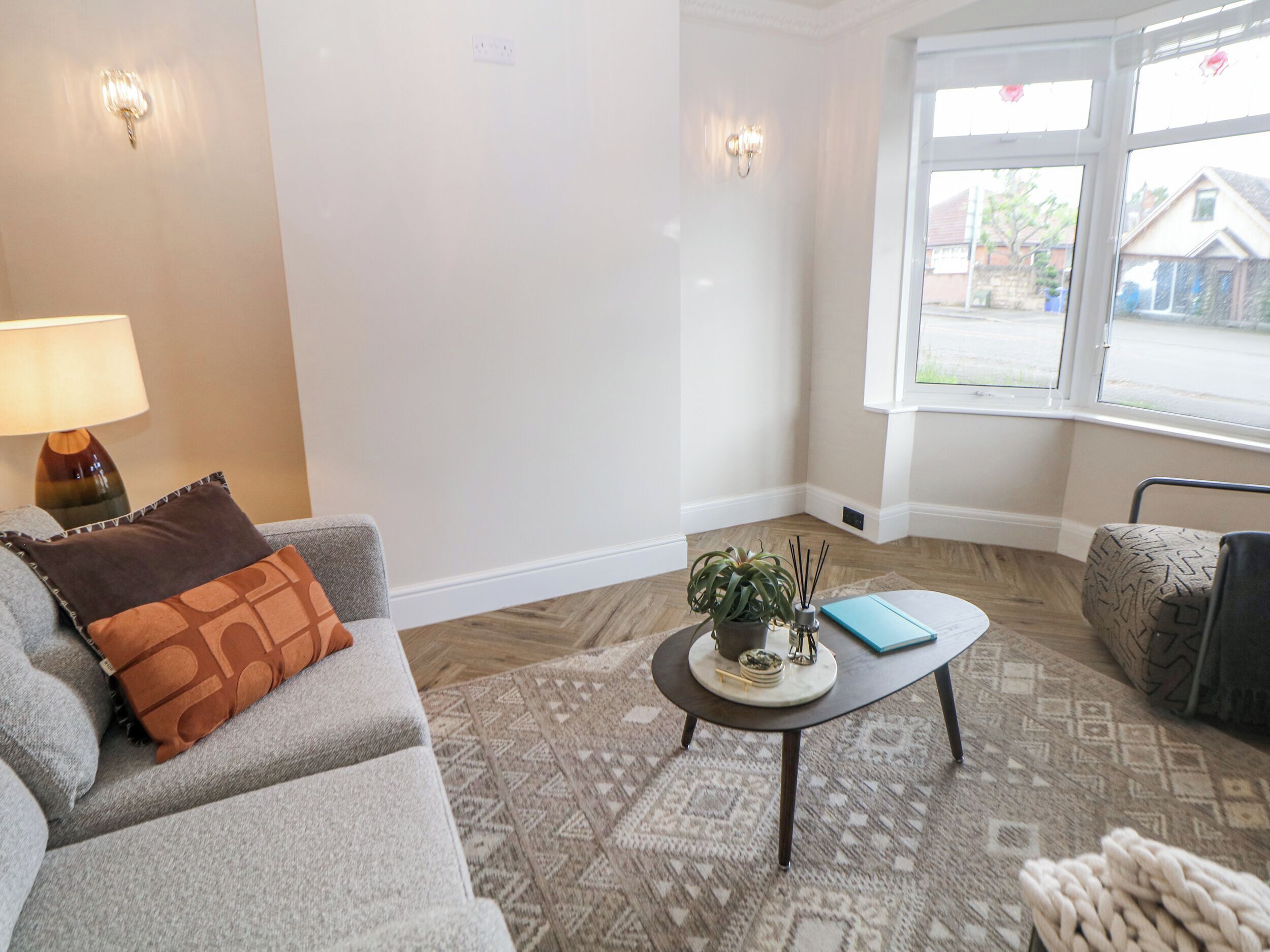 Ein Wohnzimmer mit grauem Sofa, einem kleinen Couchtisch mit Pflanze und Notizbuch, einer Lampe und einem Fenster mit Blick auf ein Haus.