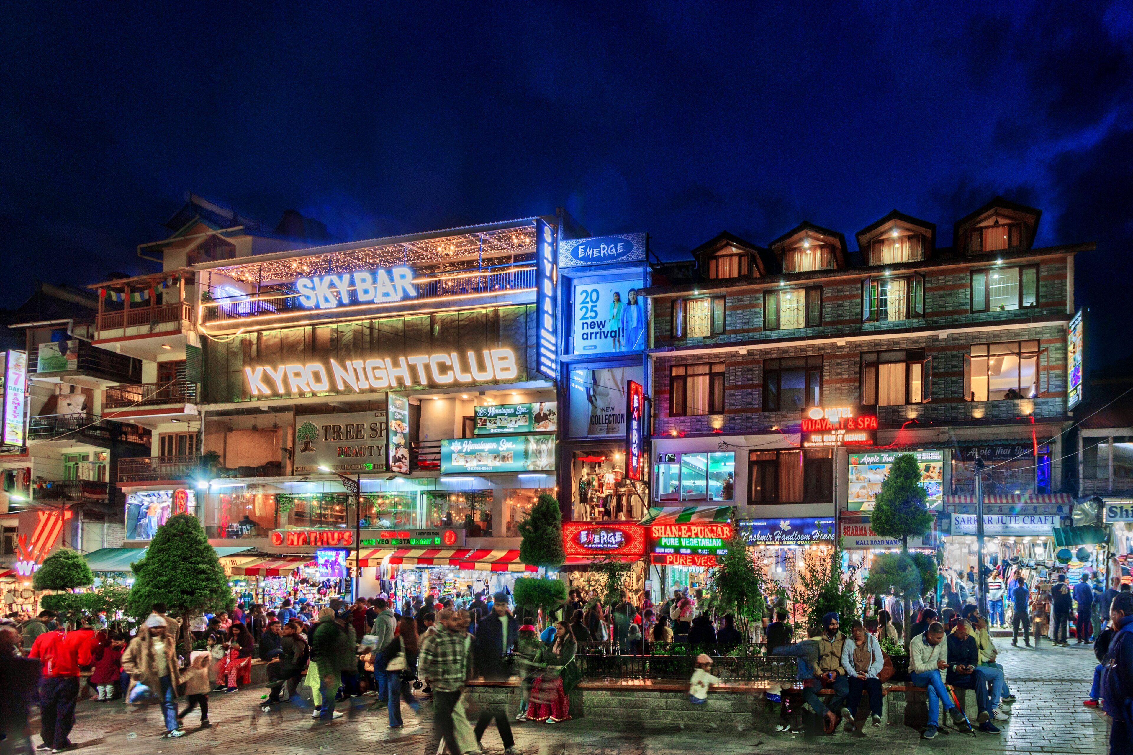 A busy urban street at night with illuminated signs for a nightclub, a bar, and various shops.