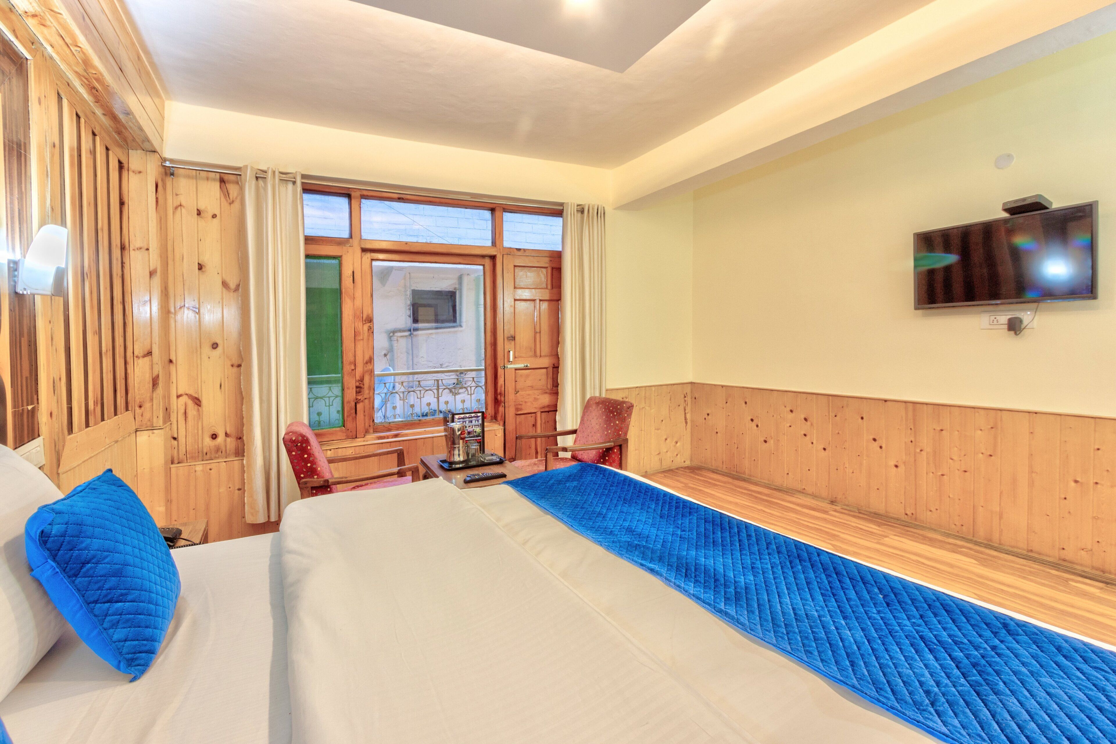 A hotel room with a large bed, wooden paneling, a television mounted on the wall, and a window with curtains.