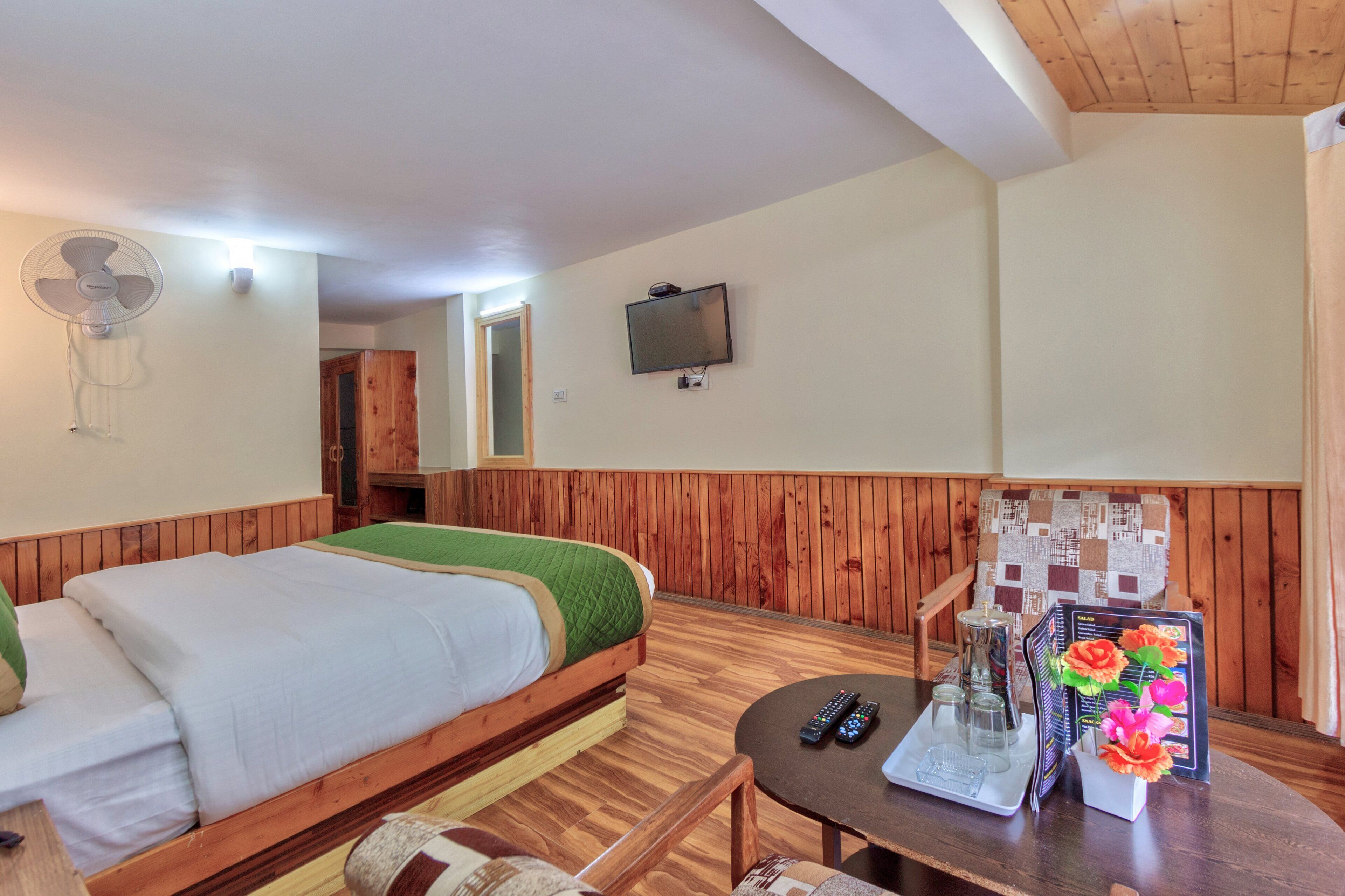 Habitación de hotel con una cama de madera, una mesita con un control remoto, un decantador de vidrio y una revista.