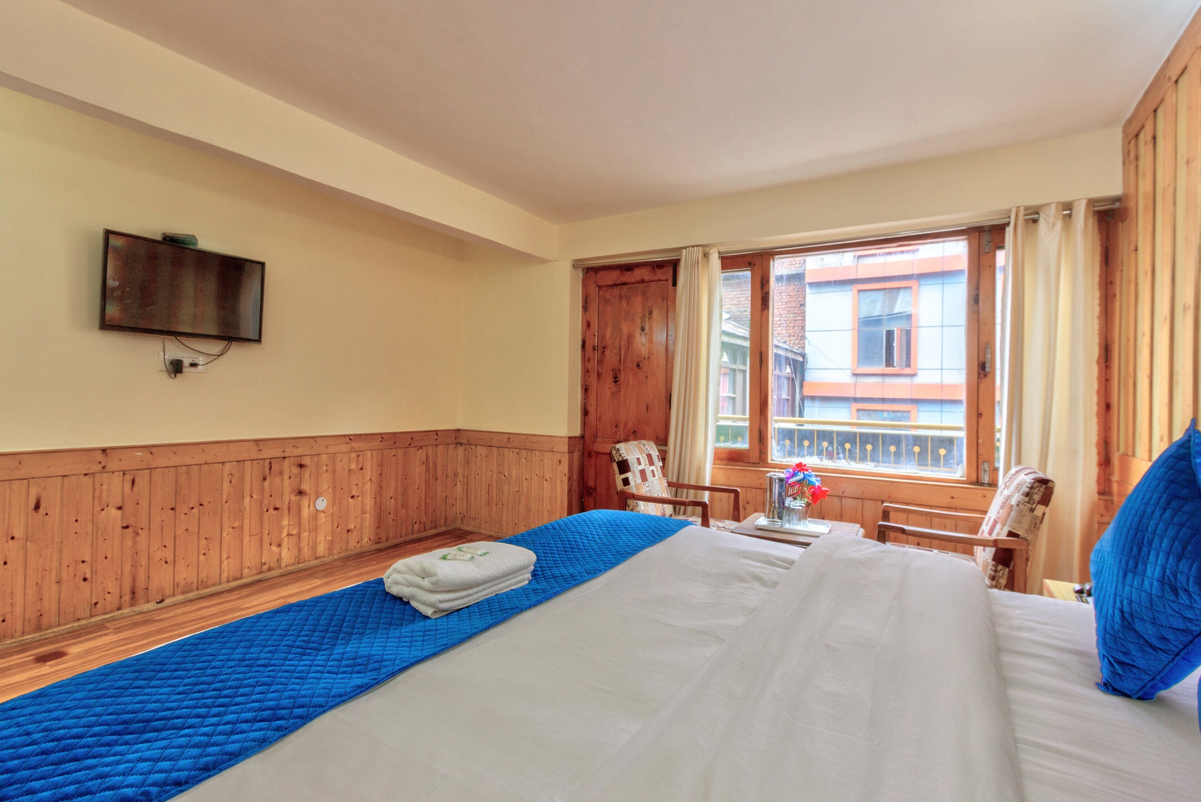 A hotel room with a large bed, wooden paneling, a television mounted on the wall, a window with curtains, and a view of a building outside.