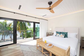 Une chambre d’hôtel avec un lit, une table à manger avec une assiette de nourriture, un ventilateur de plafond et une vue sur les palmiers, visible à travers les portes du balcon.