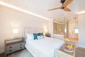 A modern bedroom with a large bed, bedside tables, a nightstand, a ceiling fan, and a bathroom visible through a glass door. - Kai Cove: Stunning Bali Beach House w/ Pool + Dock on Bio Bay (Cayman Kai)