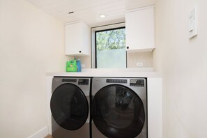 A laundry area with two front-loading washers and dryers, a window above the dryers, and a small cabinet on the wall. - Kai Cove: Stunning Bali Beach House w/ Pool + Dock on Bio Bay (Cayman Kai)