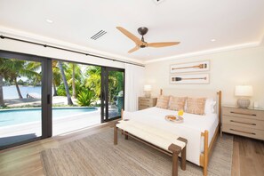 Un dormitorio moderno con una cama grande, un ventilador de techo, vistas a una piscina y palmeras, y una mesita de noche con una lámpara.