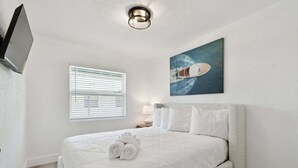 Una cama bien hecha con sábanas blancas y toallas enrolladas, un cuadro de un barco en la pared, una lámpara en el techo y una ventana con persianas.