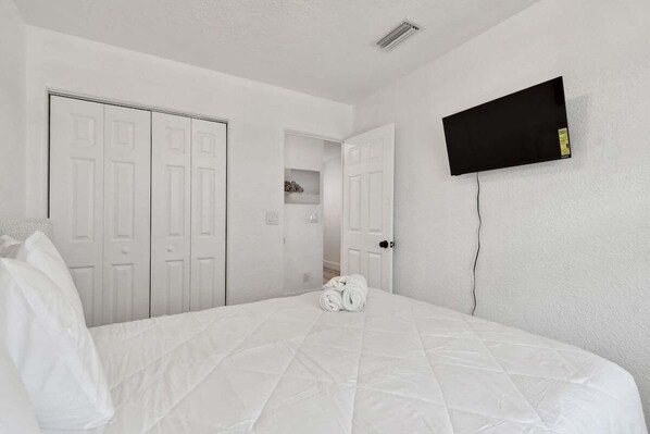 Quarto branco com uma cama, uma TV fixada na parede e um conjunto de portas brancas.
