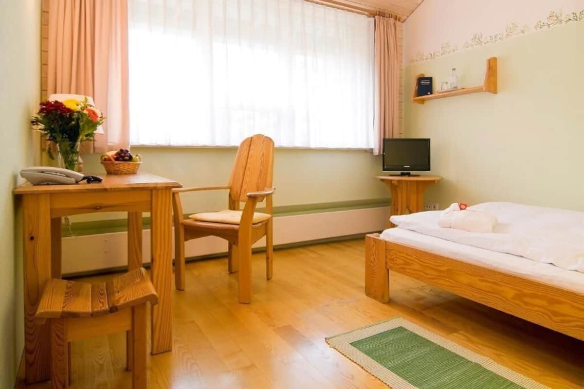 Une chambre avec un lit en bois, un bureau avec un téléphone, une chaise, un téléviseur posé sur une petite table et une fenêtre avec des rideaux.