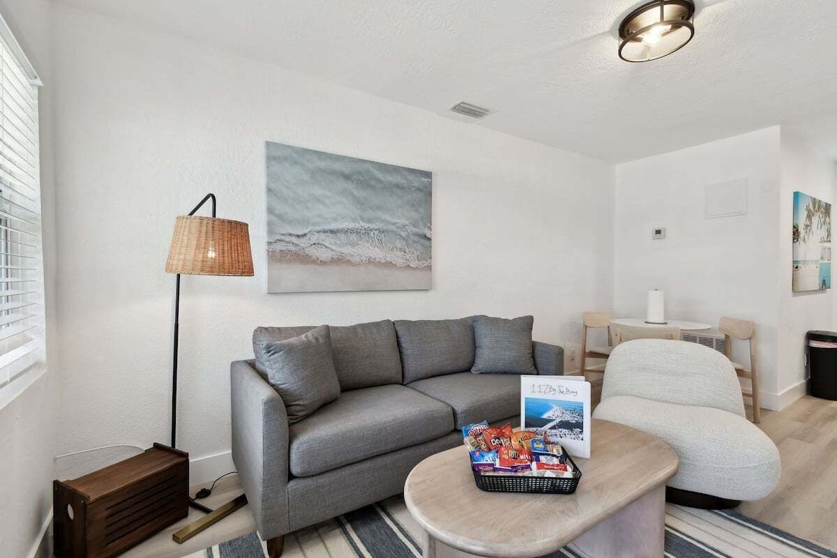 A modern living room with a grey sofa, a white armchair, a small round table with snacks, a lamp, and a beach-themed painting on the wall.