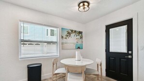 Une pièce avec une table ronde, quatre chaises, une poubelle noire, une porte noire, une fenêtre avec des stores et un tableau accroché au mur.