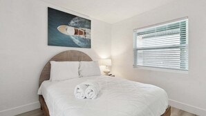 Un lit bien fait, avec des serviettes pliées, un tableau représentant un bateau accroché au mur et une fenêtre avec des stores.