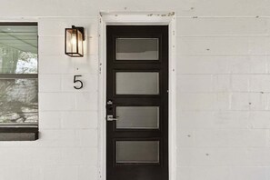 A black door with three glass panels, set within a white brick wall, features a wall-mounted light fixture above.