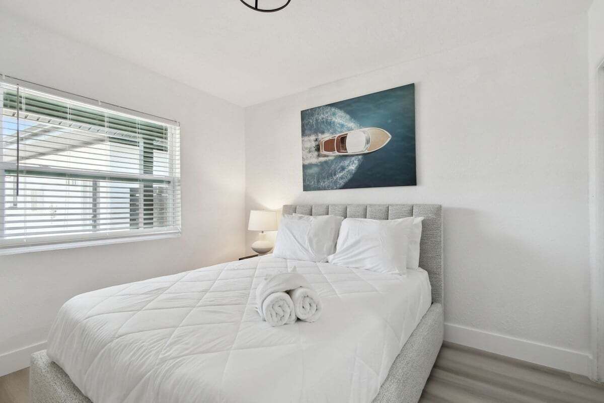 A neatly made bed with white linens and rolled towels, a grey upholstered headboard, and a painting of a boat on the wall.
