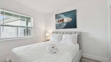 A neatly made bed with white linens and rolled towels, a grey upholstered headboard, and a painting of a boat on the wall.