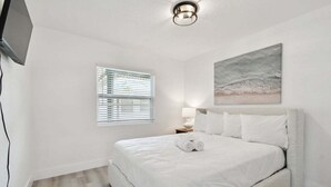 A bedroom with a bed, a window with blinds, a painting on the wall, and a lamp on a bedside table.