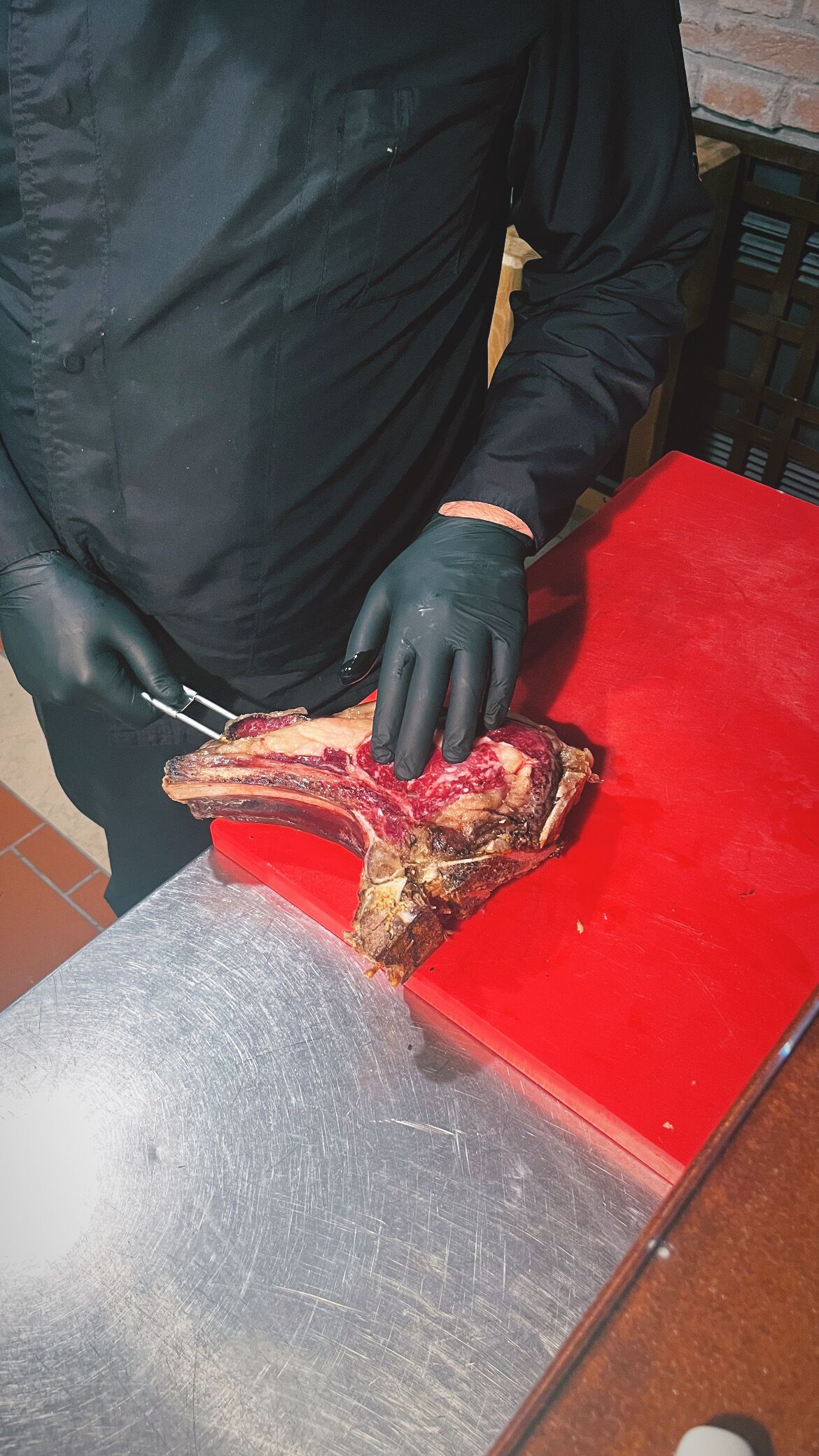 Una persona con guantes negros manipulando un gran trozo de carne sobre una tabla de cortar roja.