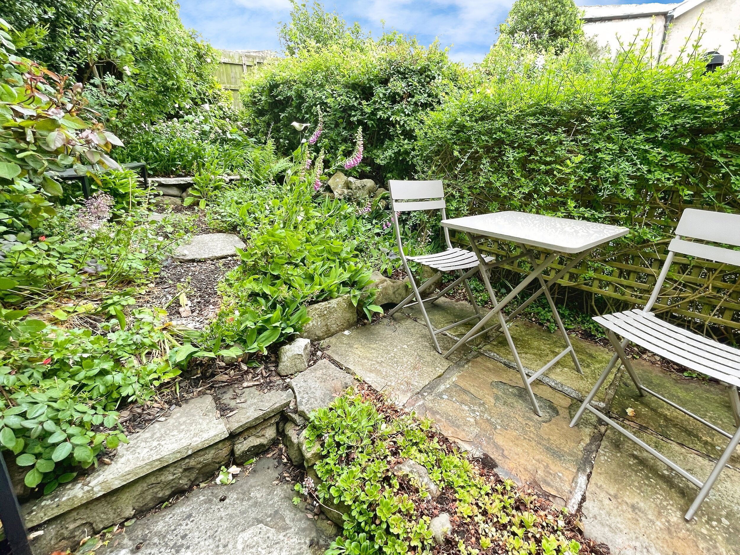 A garden patio with a table and chairs, surrounded by lush greenery and stone pathways.