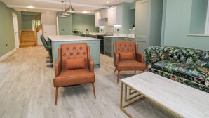 Un amplio salón con un sofá, dos sillones y una mesa de centro. Se observa una cocina con una isla central y electrodomésticos modernos al fondo.
