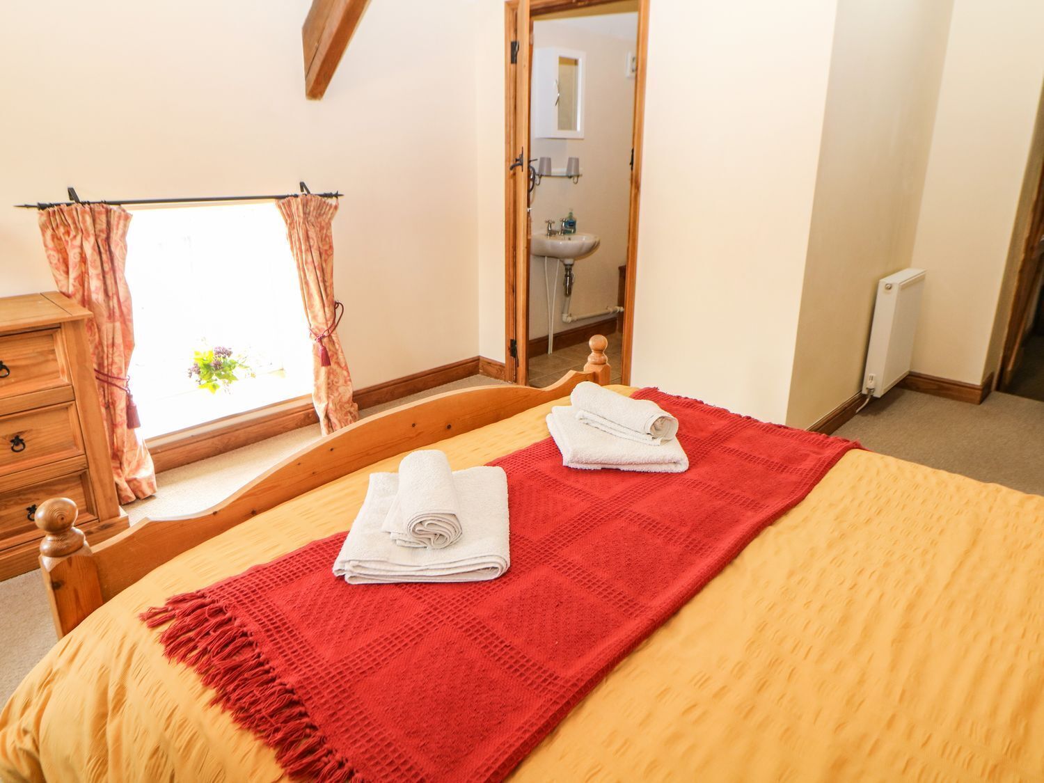 Une chambre à coucher avec un lit, une commode en bois, une fenêtre avec des rideaux et une salle de bain visible à travers une porte ouverte.