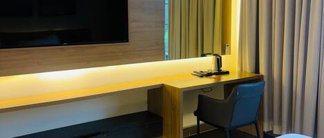 A hotel room with a bed, a desk with a chair, a flat-screen TV mounted on the wall, and an air conditioning unit.