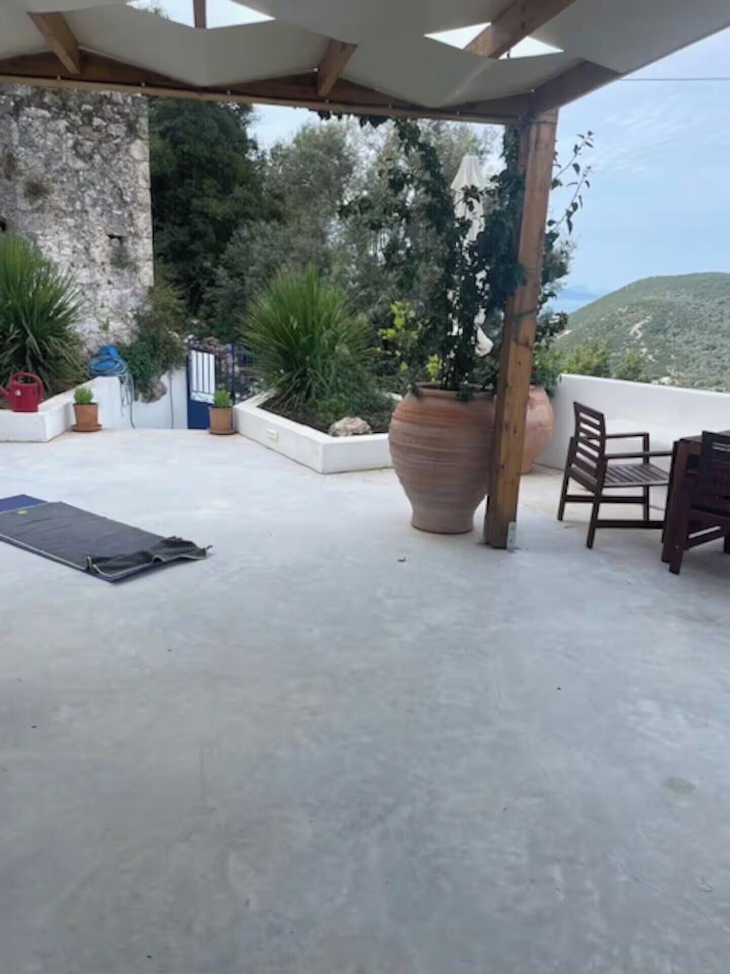 Un espace extérieur abrité, doté d’un sol en béton, de plantes en pot, d’une table et de chaises en bois, et offrant une vue sur un paysage montagneux.