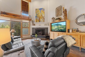 Una sala de estar moderna con chimenea, sofás de cuero, televisor y un ventanal con vistas a un área boscosa.