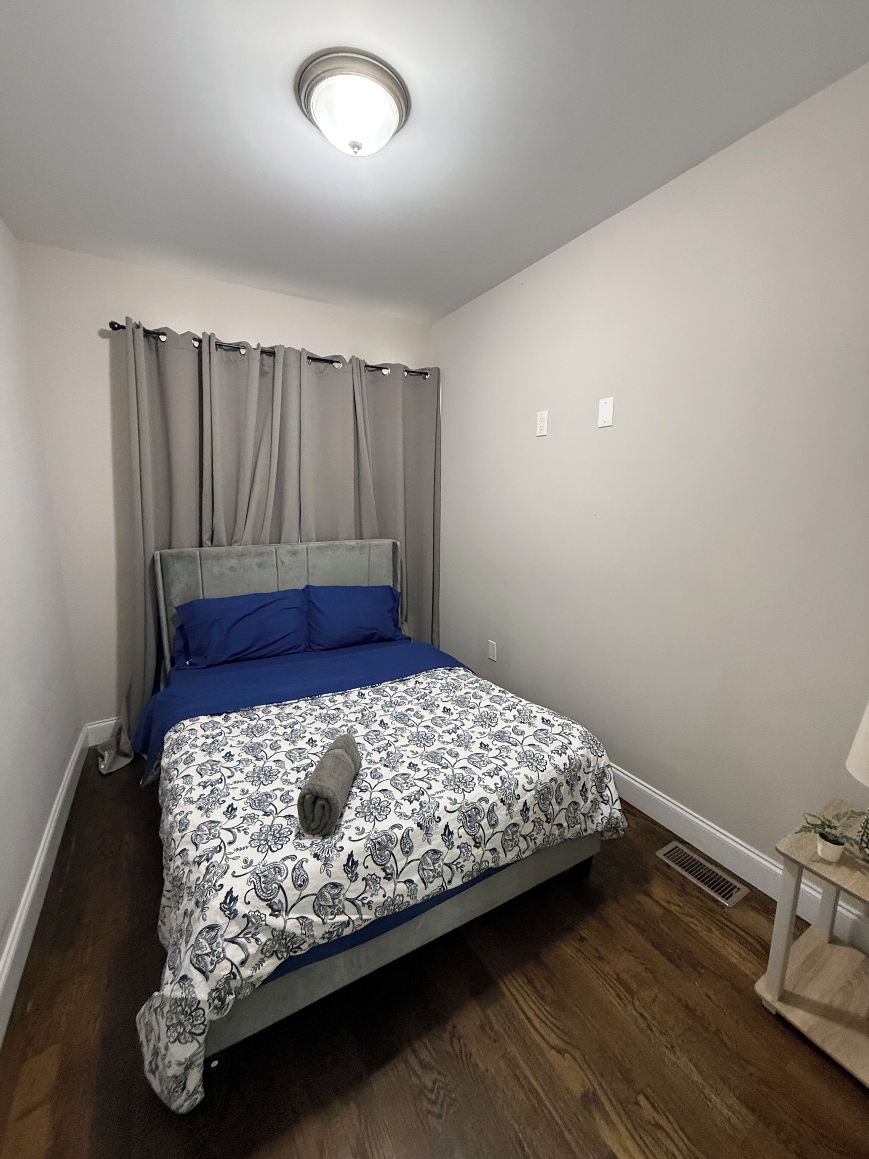 A bedroom with a bed, grey curtains, a ceiling light, a small table with a plant, and a vent.