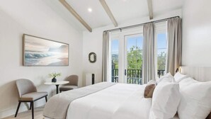 A bedroom with a large bed, a desk, and a chair. There is a window with curtains and a view of greenery outside. - Casa Riviera 21 - Boutique Suites (Santa Barbara)