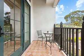 A balcony with a checkered floor, two metal chairs, a small round table, and a glass door leading to an outdoor area with a view of trees and a fence.