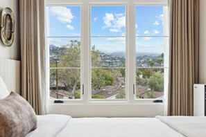A bedroom with a large window offering a view of a residential area and mountains.