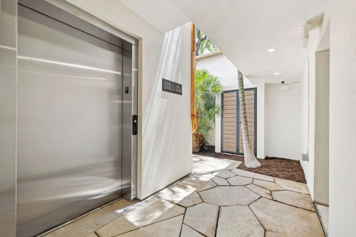 A modern hallway with a closed elevator, a sign indicating levels, and a stone-tiled floor leading to a gated entrance.